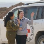 אגם ברגר בסווטשירט אפור מנופפת בחיוך לצד קצינה במדים, ליד רכב של הצלב האדום, ביום שחרורה מהשבי