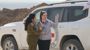 אגם ברגר בסווטשירט אפור מנופפת בחיוך לצד קצינה במדים, ליד רכב של הצלב האדום, ביום שחרורה מהשבי