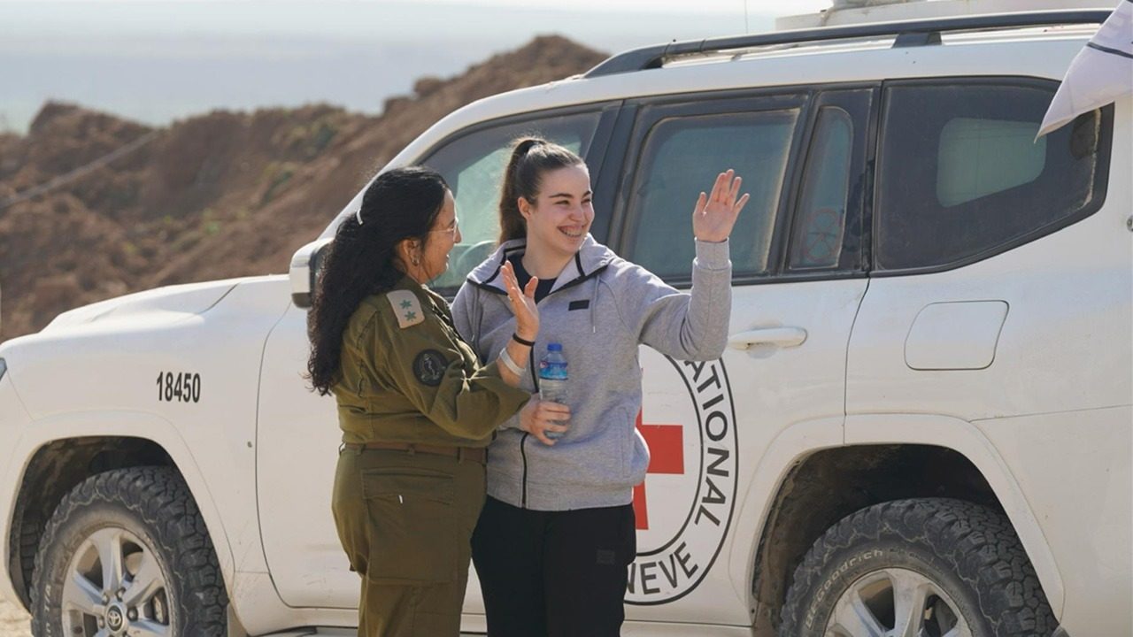 אגם ברגר בסווטשירט אפור מנופפת בחיוך לצד קצינה במדים, ליד רכב של הצלב האדום, ביום שחרורה מהשבי