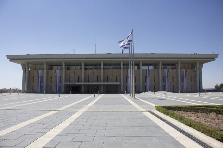 בניין הכנסת בירושלים מהכיכר הקדמית, עם שלושה דגלי ישראל מתחת לשמיים כחולים