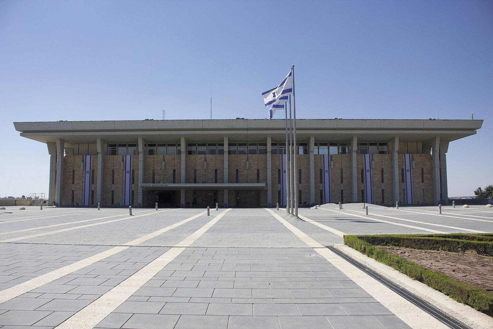 בניין הכנסת בירושלים מהכיכר הקדמית, עם שלושה דגלי ישראל מתחת לשמיים כחולים