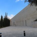 בניין בית המשפט העליון בירושלים: קיר אבן ארוך, חצר מרוצפת ושורת ברושים מתחת לשמיים כחולים