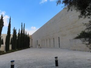 בניין בית המשפט העליון בירושלים: קיר אבן ארוך, חצר מרוצפת ושורת ברושים מתחת לשמיים כחולים
